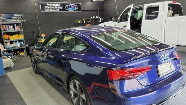 Blue car being tinted in a garage.  A white truck is nearby. Tools and supplies are visible.