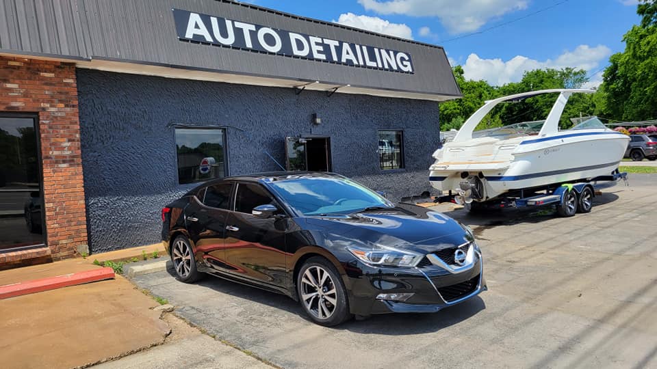 Black car in front of auto detailing shop; boat on trailer beside building.