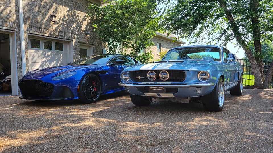 Blue Aston Martin and classic Shelby Mustang parked on a gravel driveway in front of a stone house.