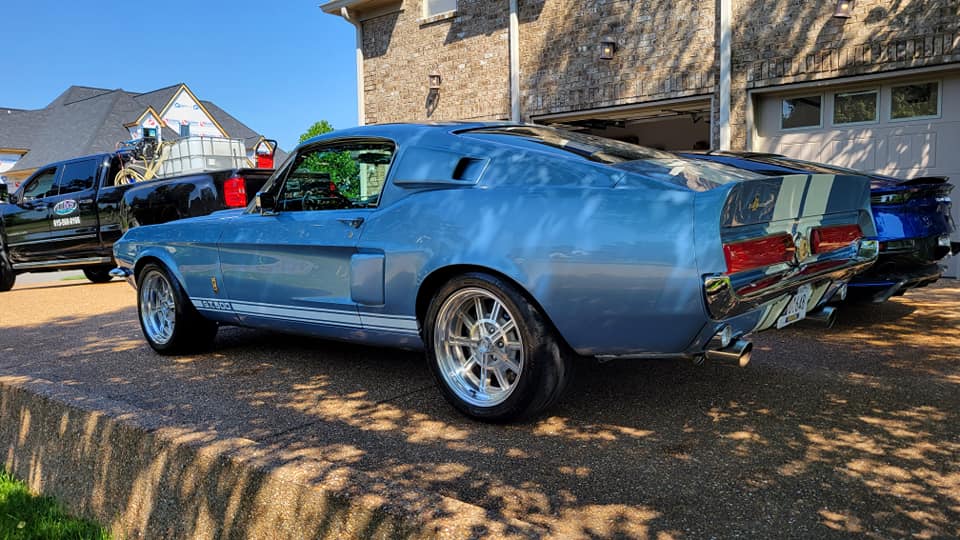 Blue classic Ford Mustang with white racing stripes parked near a house.