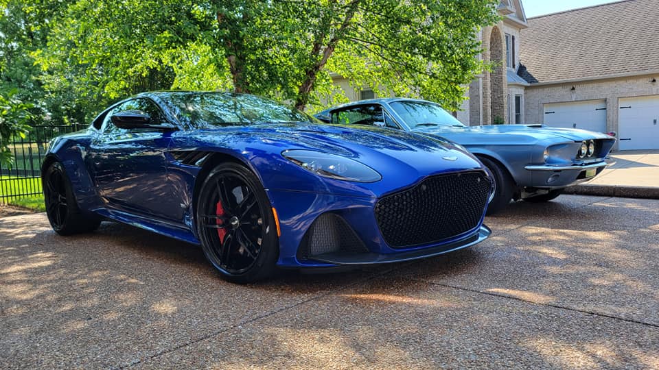 Blue Aston Martin and a classic blue Mustang parked on a paved driveway.