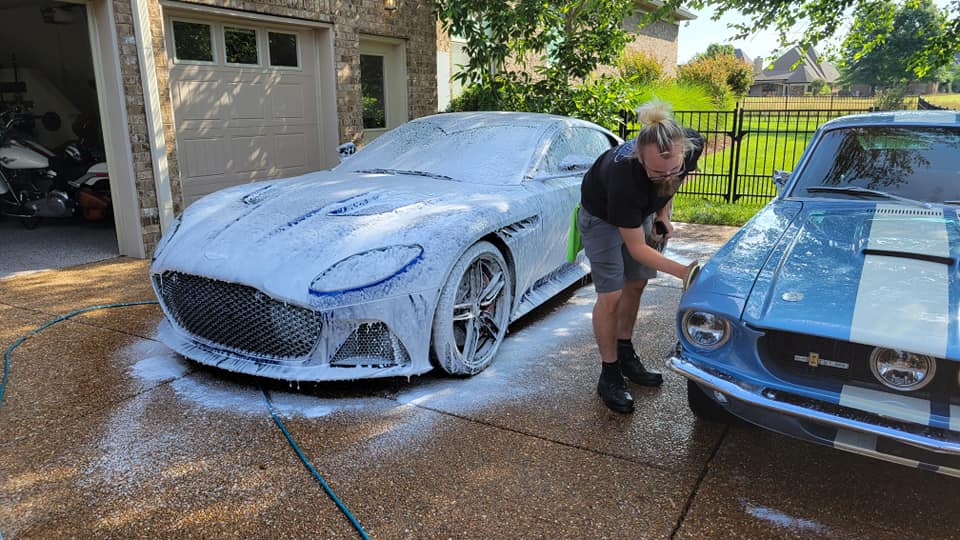 Man washing a white Aston Martin and a blue Mustang in a driveway.