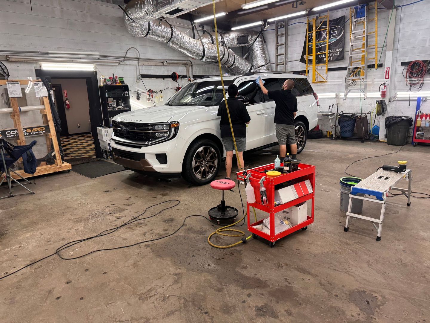 Two people tinting windows of a white SUV inside a garage; tools and equipment surround the vehicle.