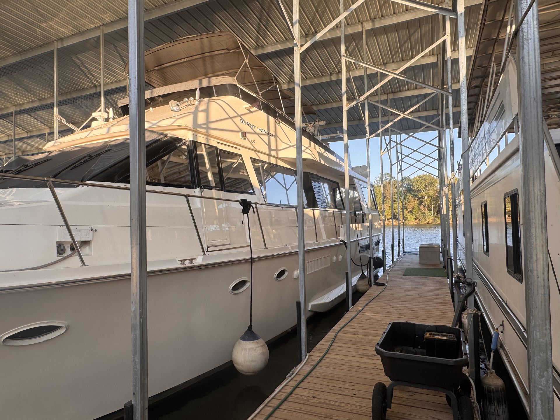 Two white boats docked under a metal-framed boat house, dock with a small utility cart, water in the distance.