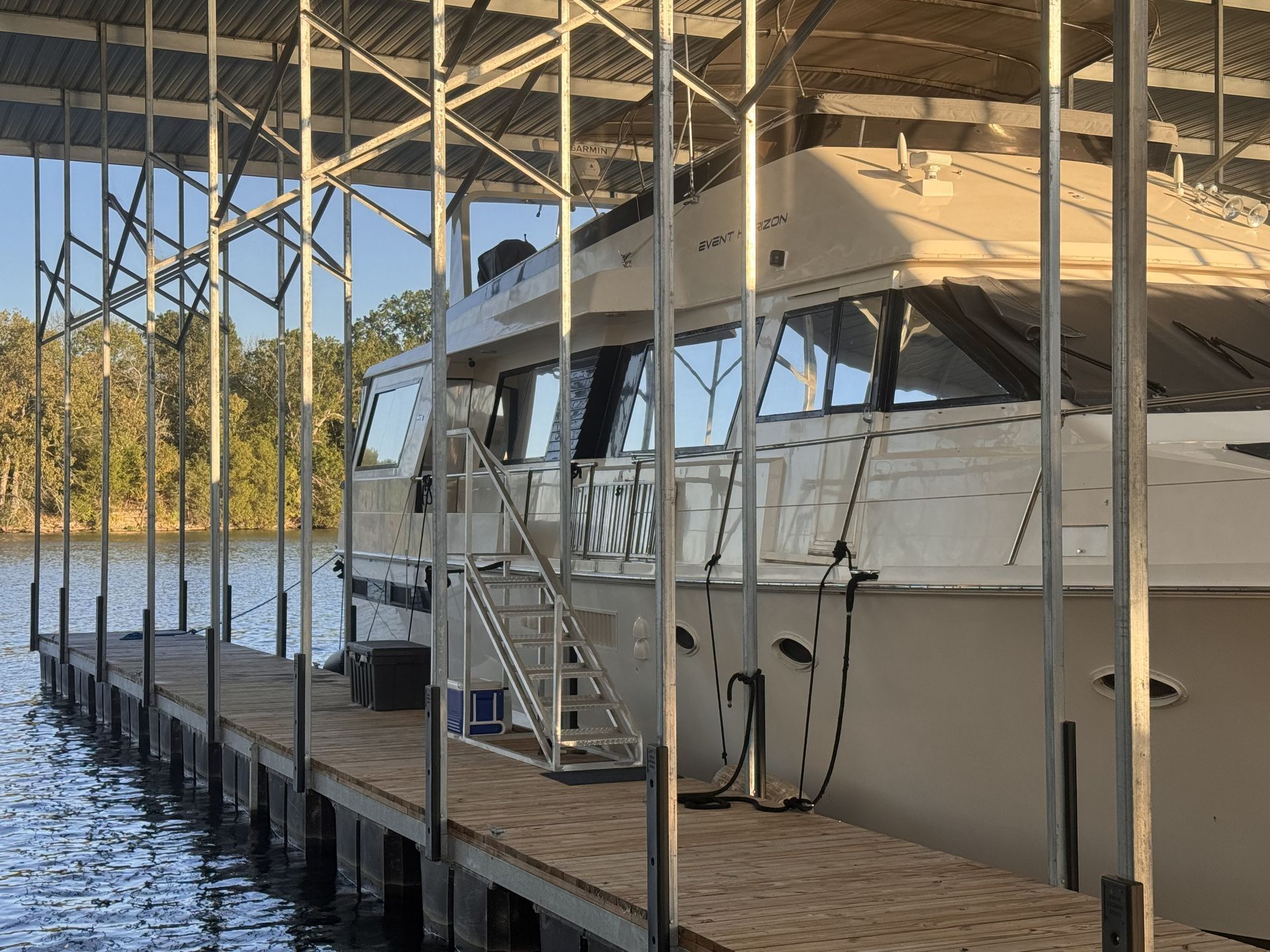 Houseboat docked at a wooden pier under a covered shelter; blue water, trees in the background.