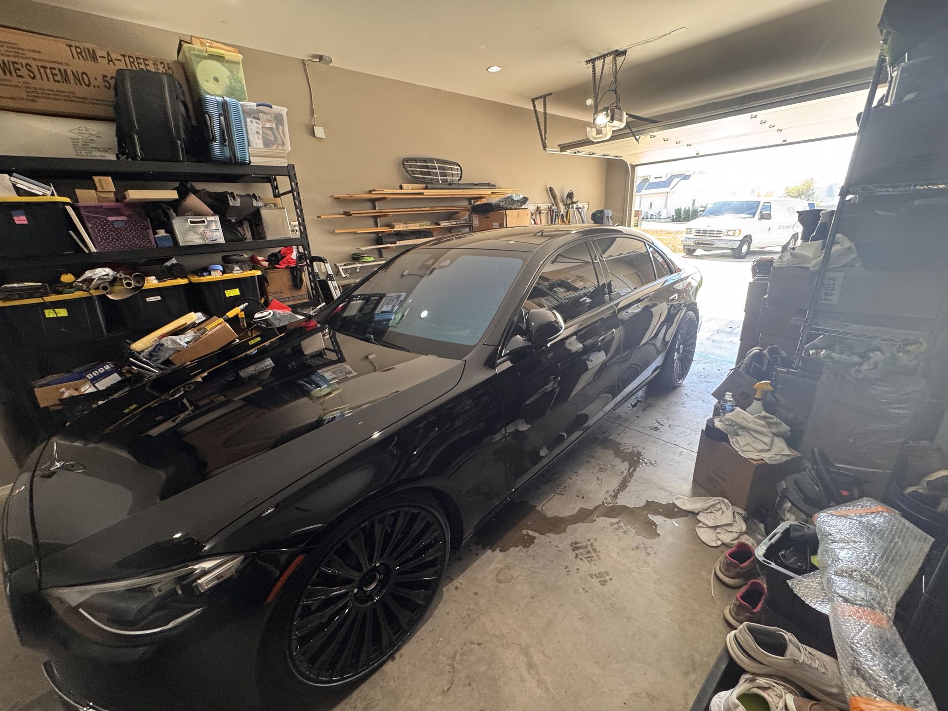 Black car parked in a cluttered garage; tools and boxes on shelves; open garage door with a car visible outside.