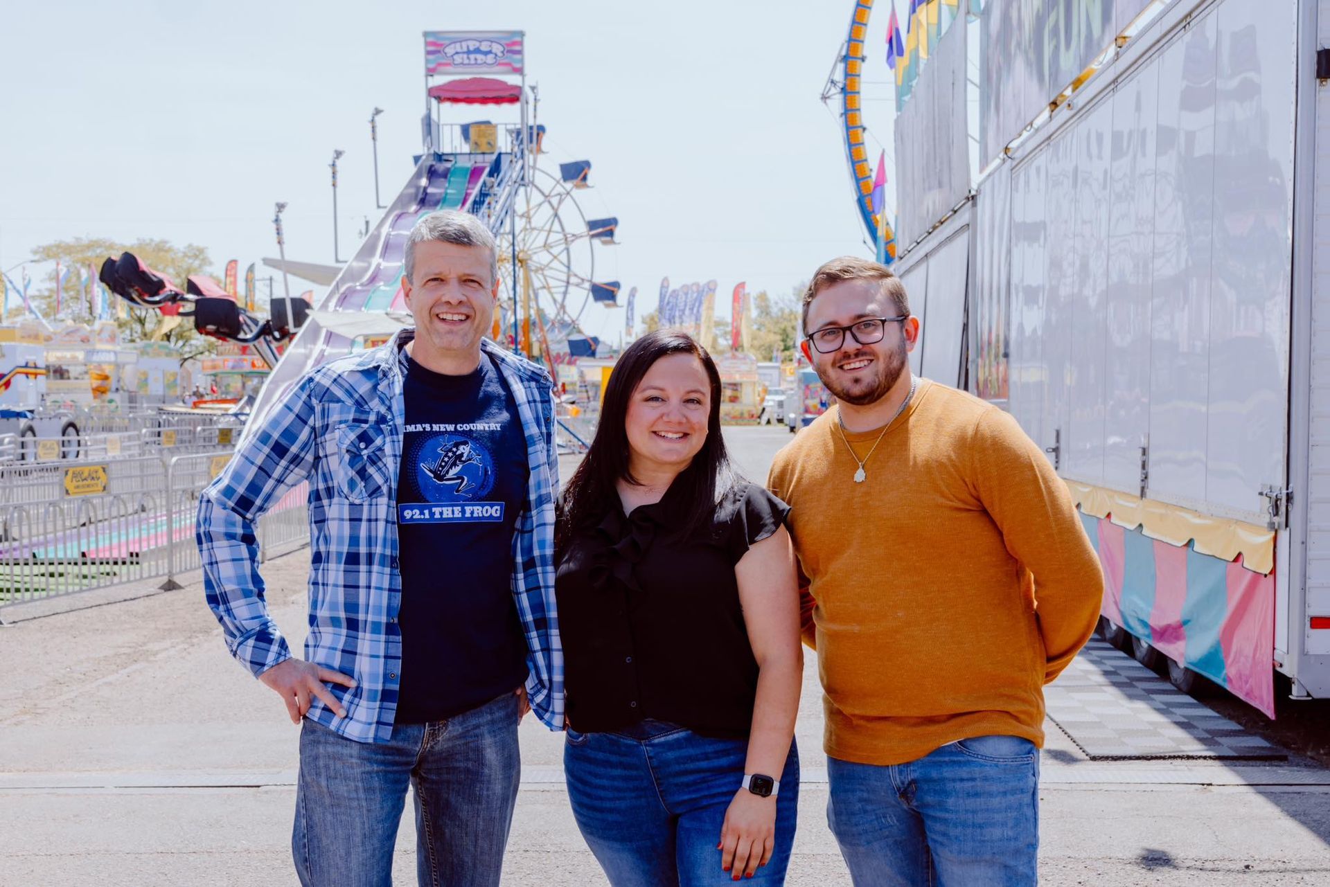 Three people stand together smiling at a carnival with rides in the background under a bright, sunny sky.