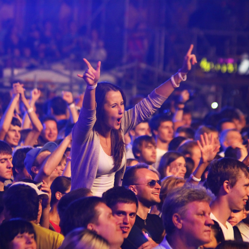 A crowd at a dimly lit, blue-tinted concert, with one person in the foreground excitedly cheering with arms raised high.