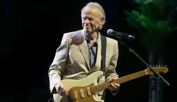Singer onstage holding a cream electric guitar beside a microphone, wearing a light blazer under stage lights