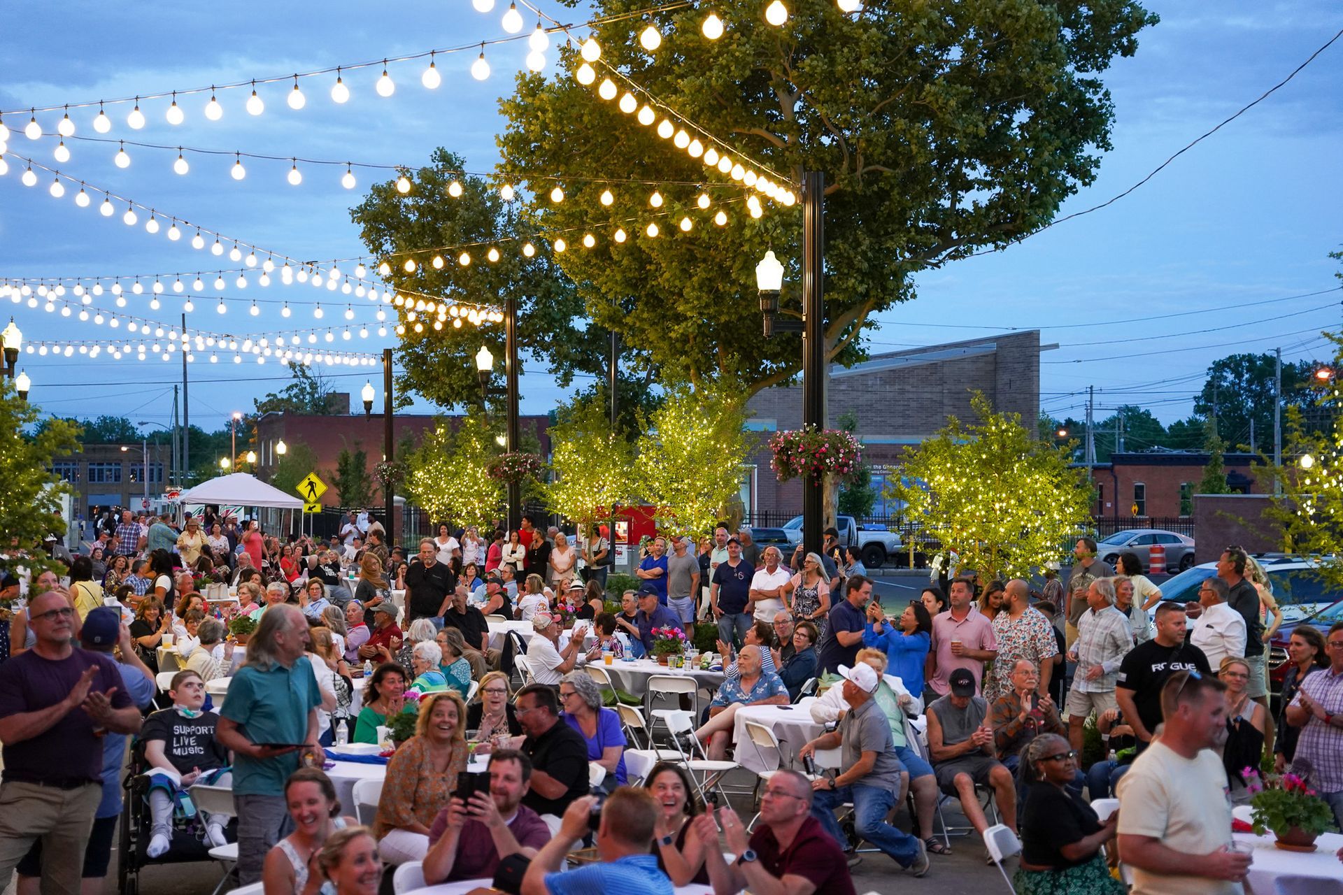 Crowd gathered outdoors under string lights at dusk, seated and standing in a lively public event