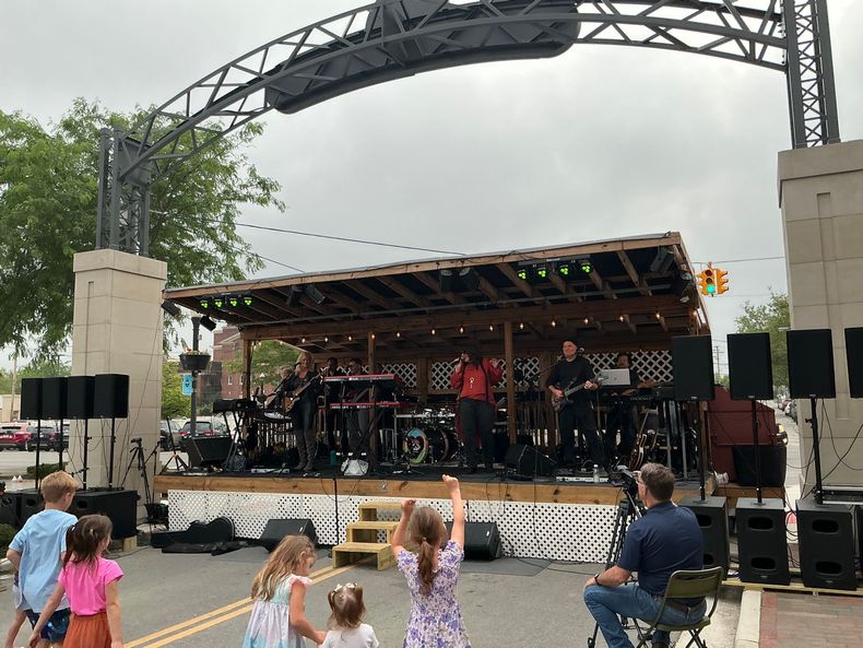 A band performs on an outdoor wooden stage under a large arched frame while several people watch from the street.