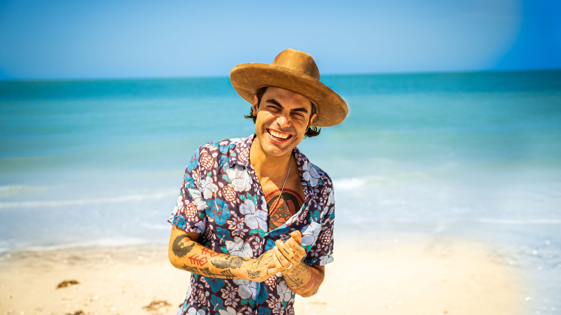 Niko Moon smiling with tattoos wearing a cowboy hat and floral shirt, standing on a sunny beach with the ocean behind.