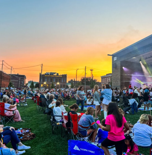 Crowd seated on a lawn at an outdoor concert at sunset, with stage lights and tents nearby.