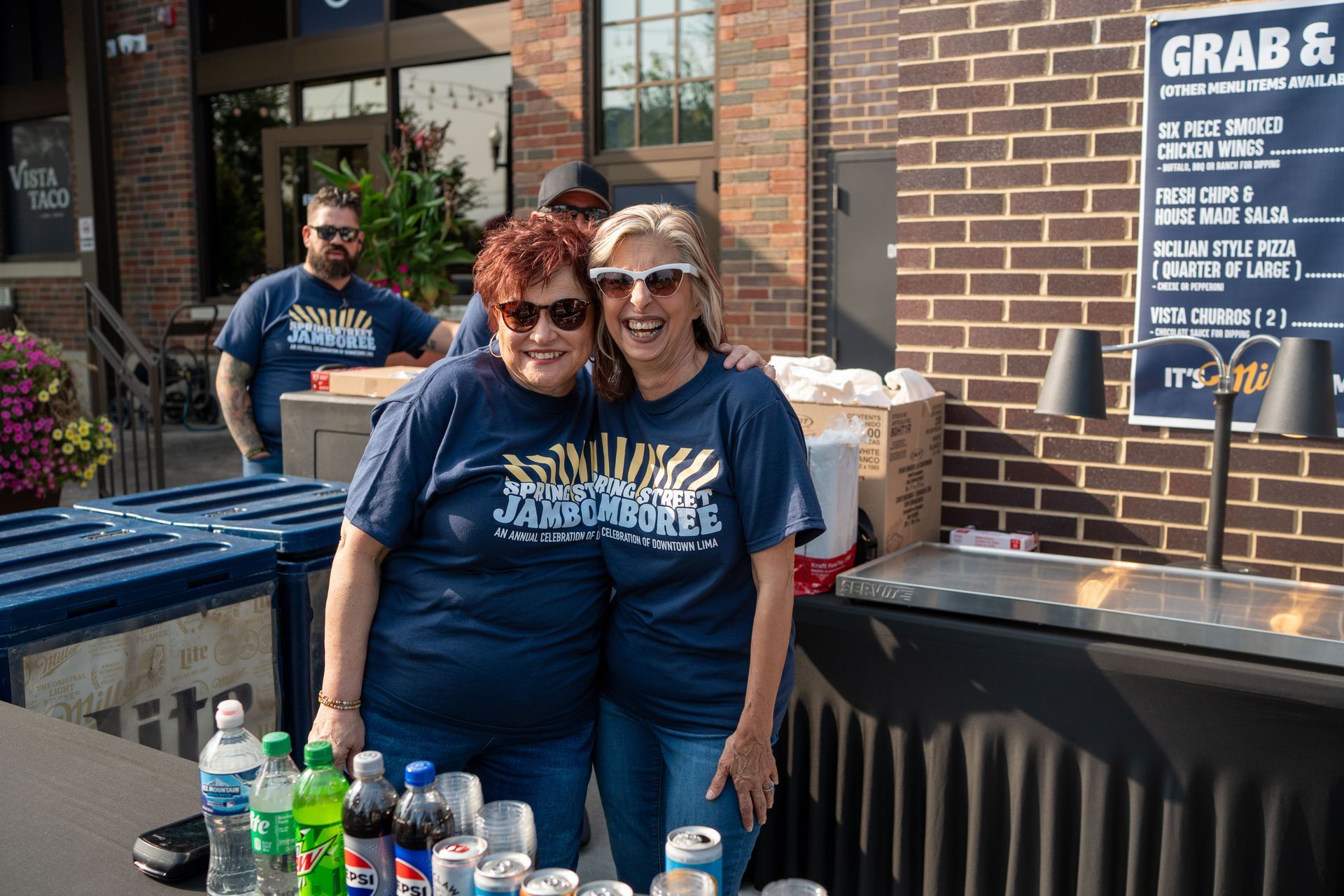 Two smiling volunteers in navy shirts standing behind a table with drinks outside a brick building
