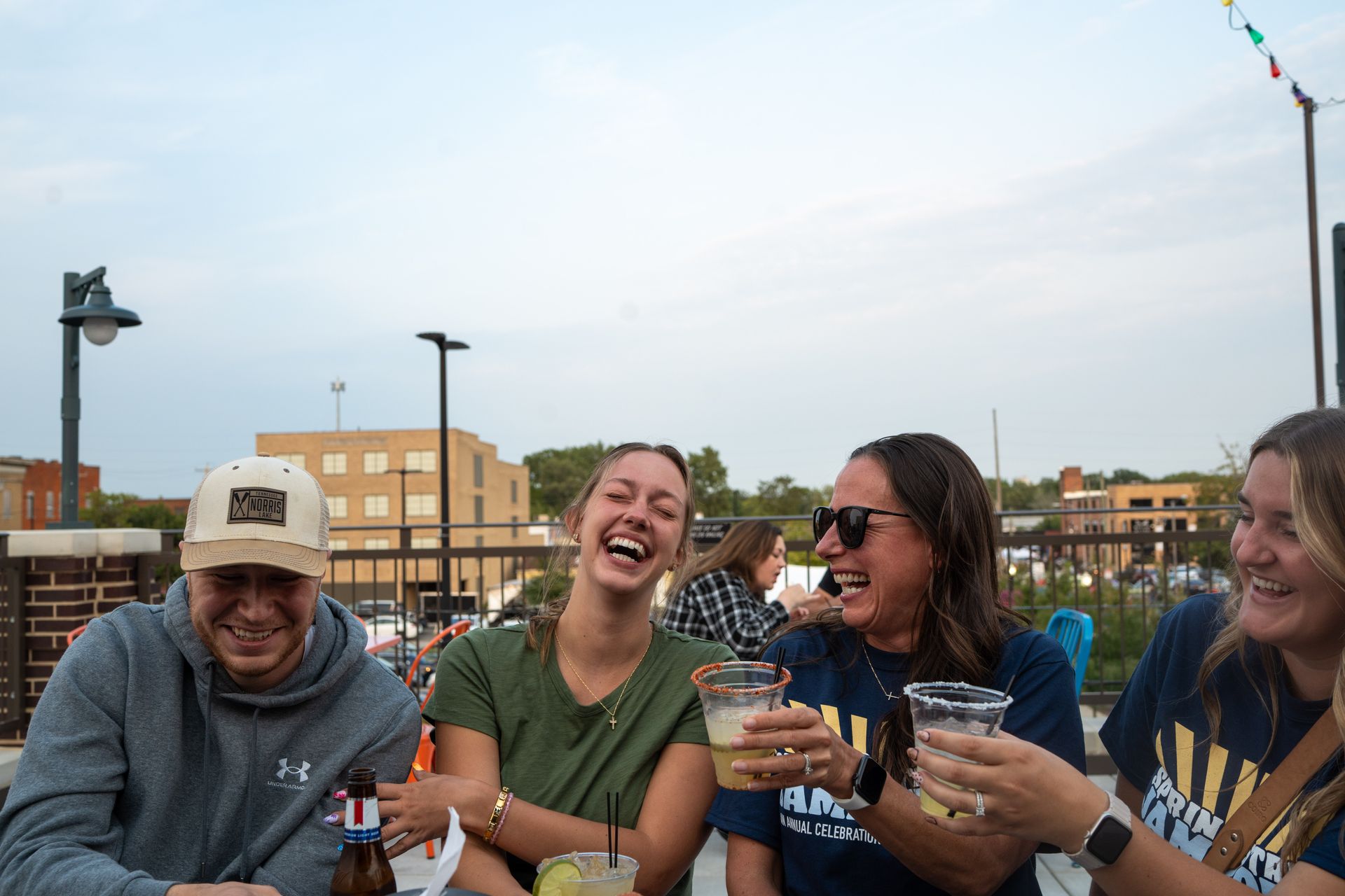 Four friends laughing and holding drinks at an outdoor table at sunset