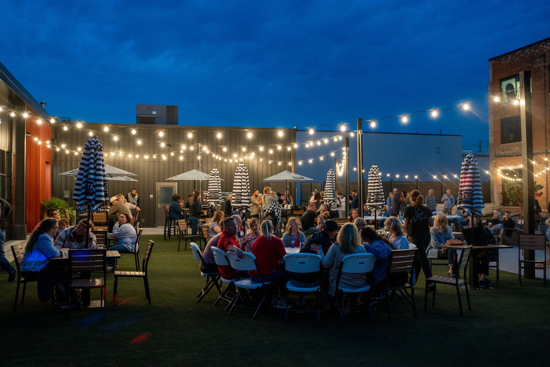 An outdoor courtyard at dusk, filled with people sitting at tables under strings of glowing lights.