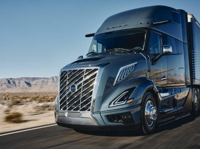 A modern, dark-gray Volvo semi-truck driving on a highway through a desert landscape under a clear blue sky.