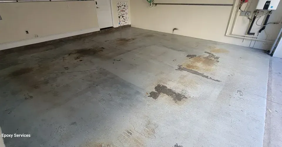 Garage interior with oil stains on a gray concrete floor, beige walls, and overhead pipes.