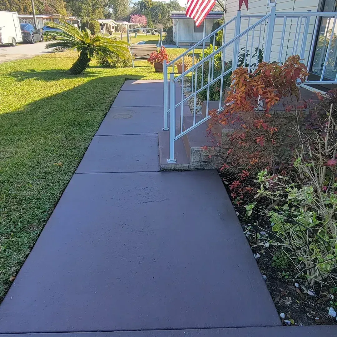 A paved walkway leading to a house with a white railing and a small American flag; greenery on both sides.