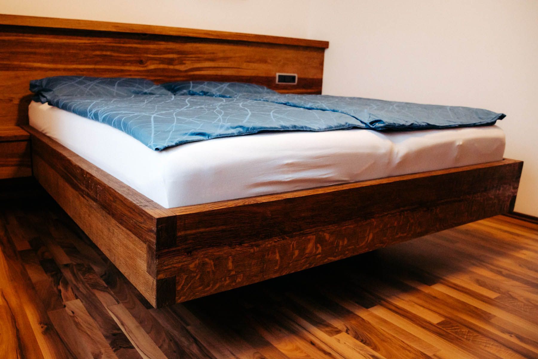 Schwebendes Holzbett mit blauer Decke und weißen Laken. Brauner Hartholzboden.