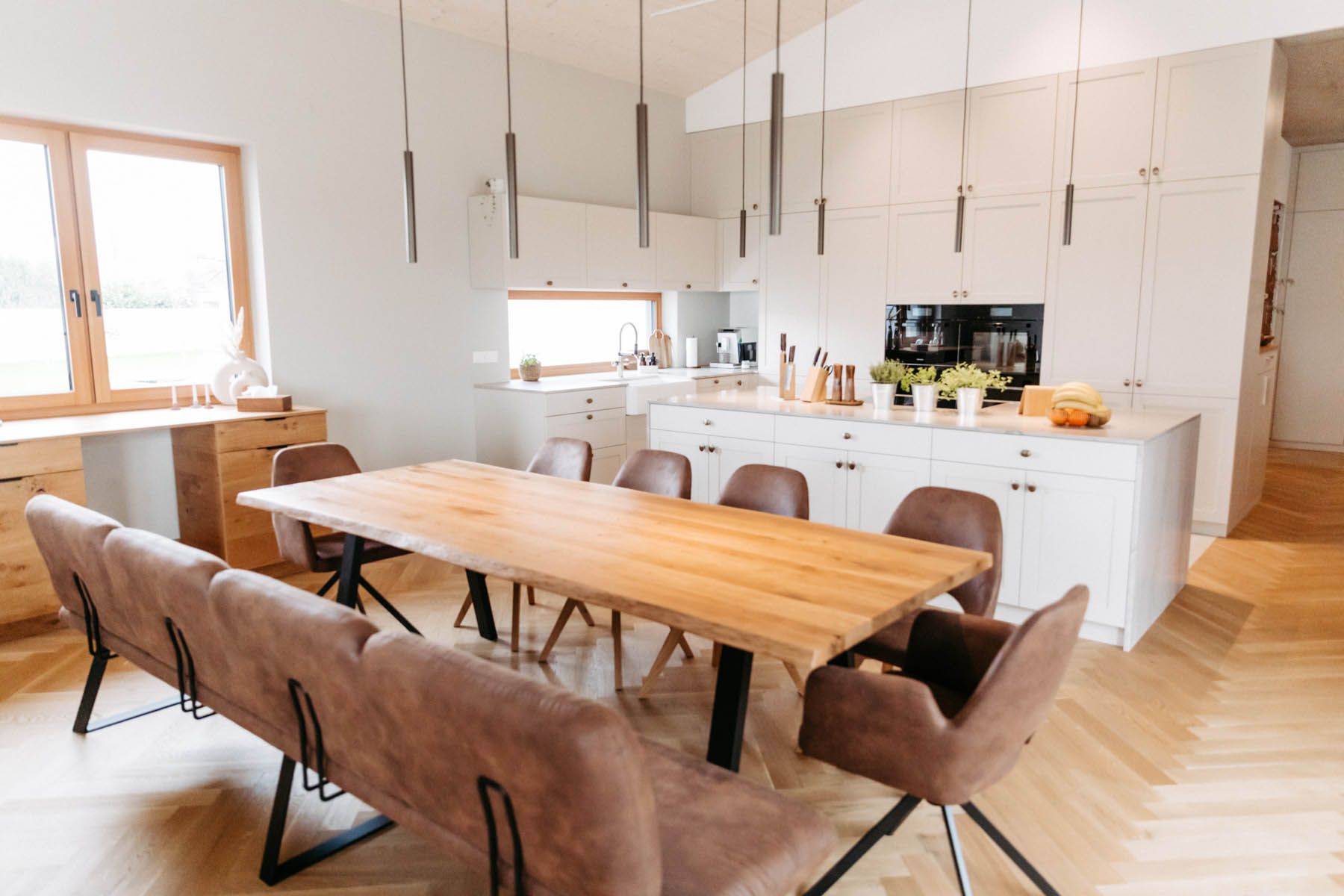 Modernes Esszimmer mit großem Holztisch, braunen Stühlen und weißer Küche.