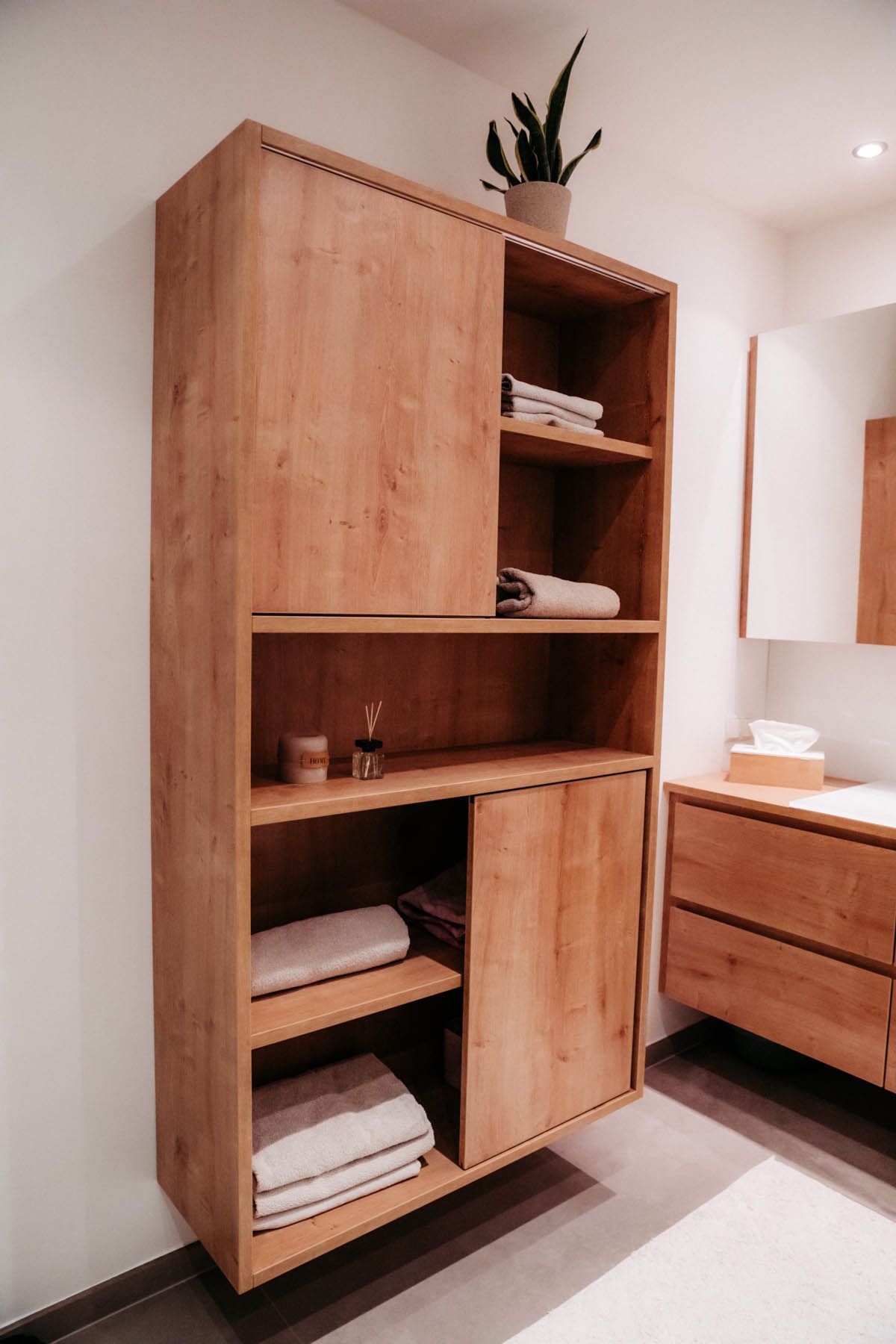 Badezimmerschrank aus Holz mit Schiebetüren und Regalen für Handtücher, Toilettenartikel und eine Pflanze.