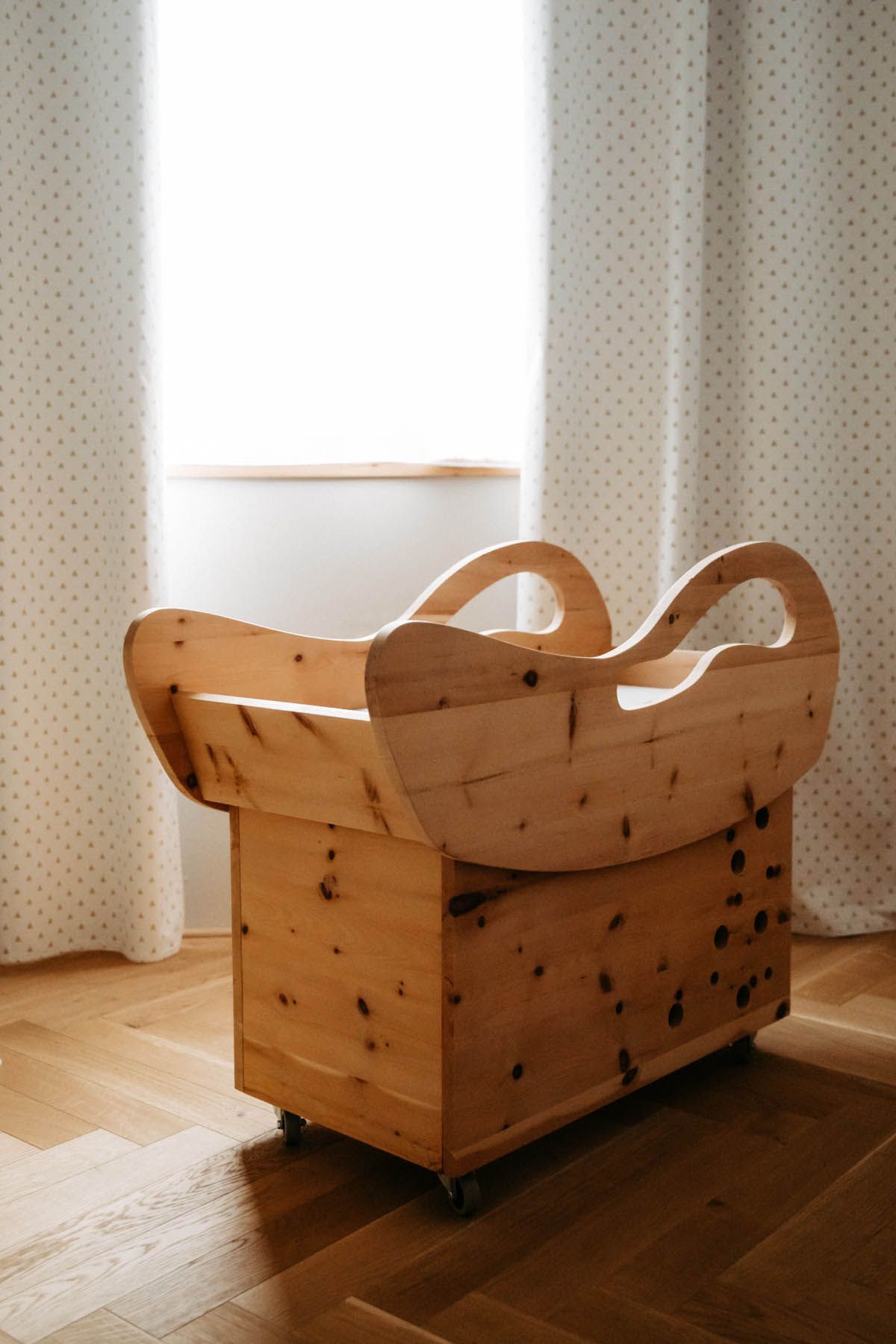 Holzspielzeugkiste mit geschwungenen Griffen auf Rädern vor einem Fenster.