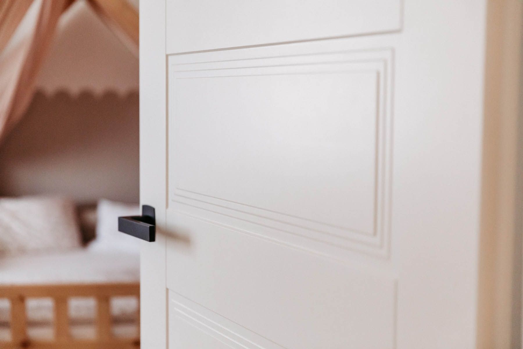 Weiße Tür mit schwarzem Griff, leicht angelehnt, gibt den Blick auf ein Kinderzimmer mit einem Bett frei.