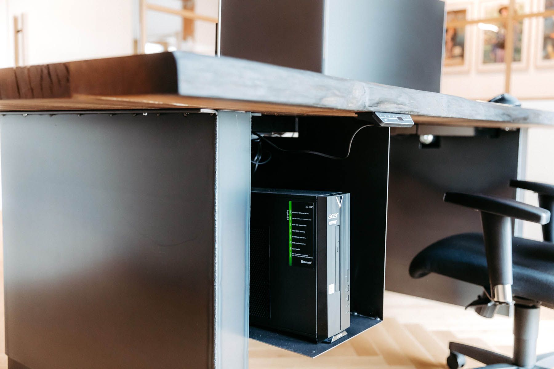 Schwarzer Computerturm unter einem Schreibtisch montiert. Der Schreibtisch ist dunkelgrau und schwarz. Im Hintergrund steht ein Bürostuhl.