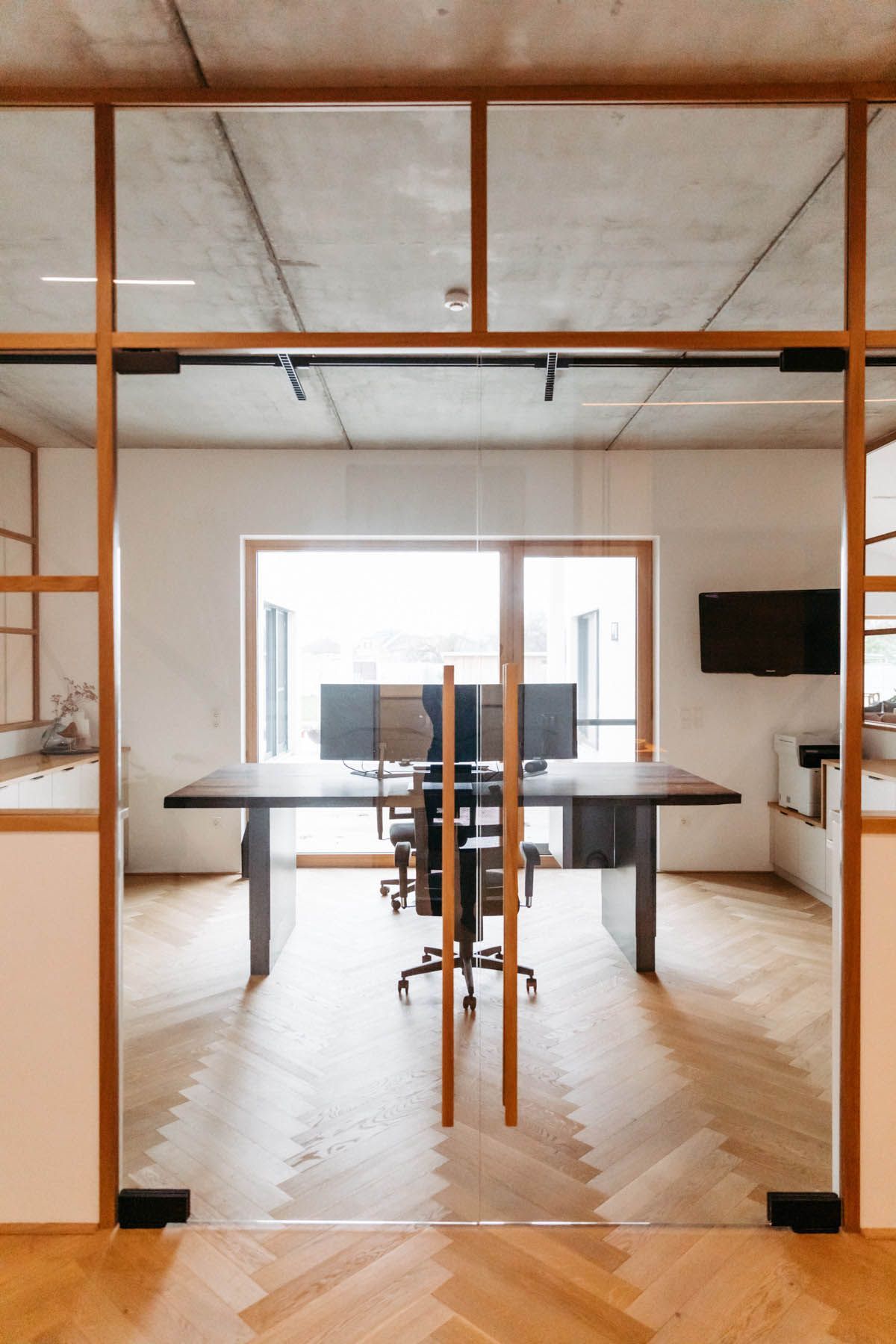 Büro mit Glaswänden, Schreibtisch, Monitoren und Stuhl. Helle Holzböden und -verkleidung, Betondecke.