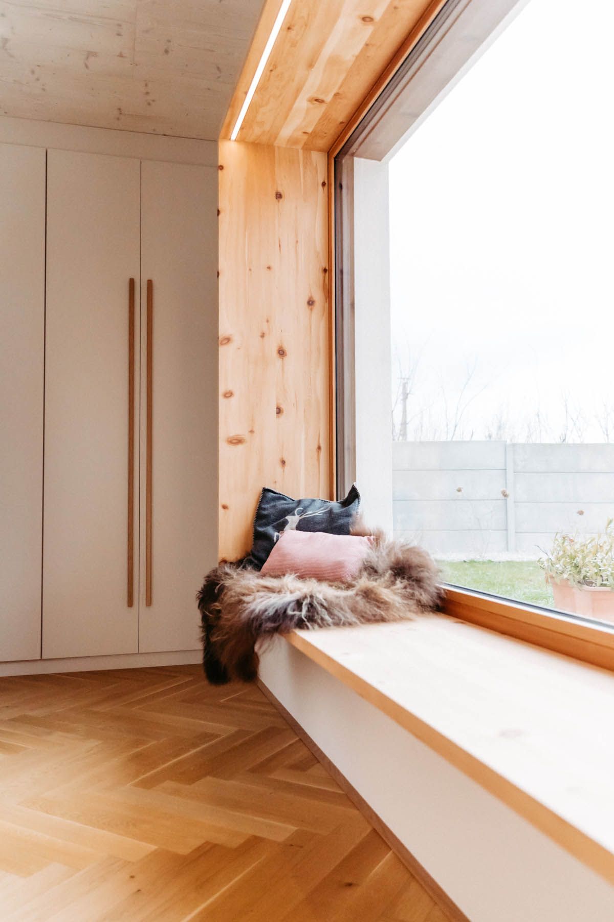 Gemütlicher Fensterplatz mit Holzverkleidung, Felldecke und Kissen, neben einem weißen Schrank mit Holzgriffen.