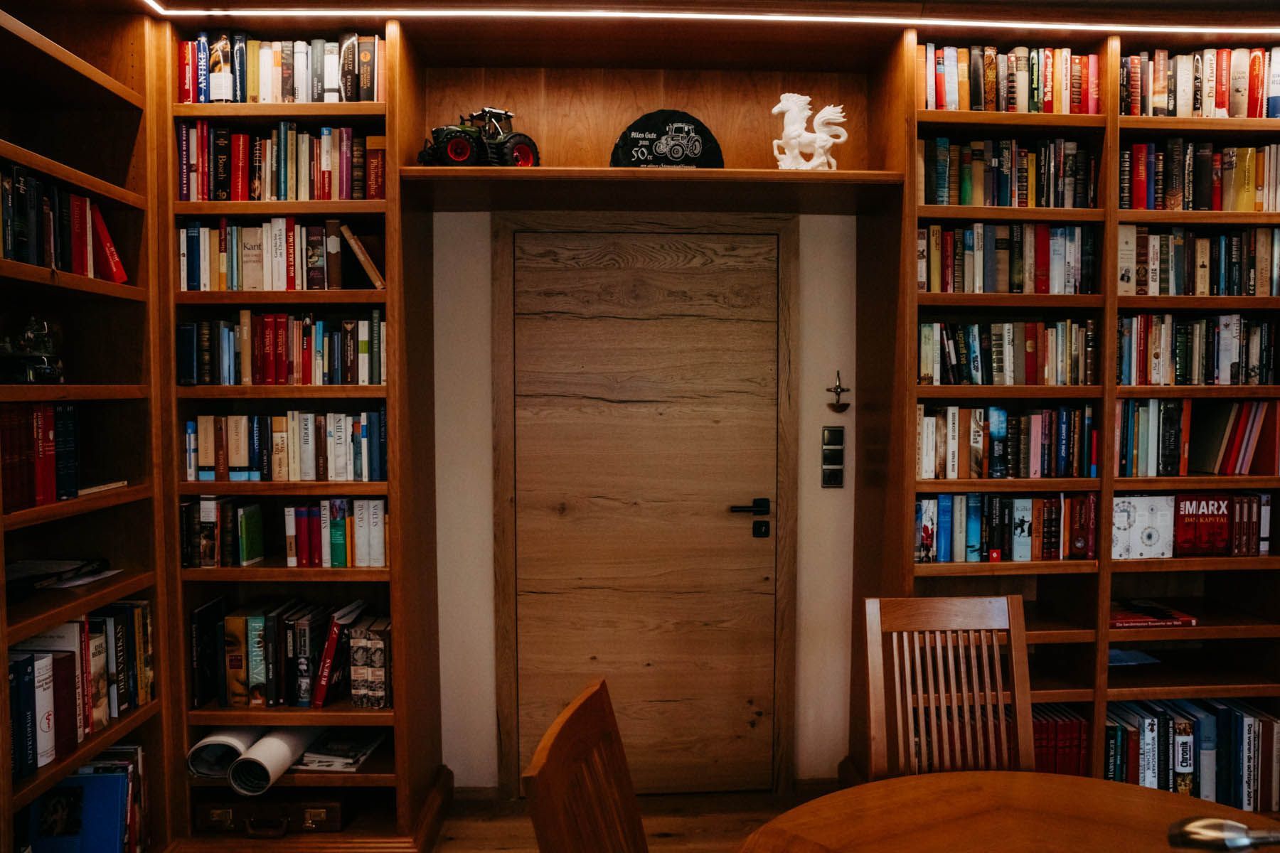 Holzbücherregale neben einer Holztür, gefüllt mit Büchern. Holztisch und Stühle im Vordergrund.