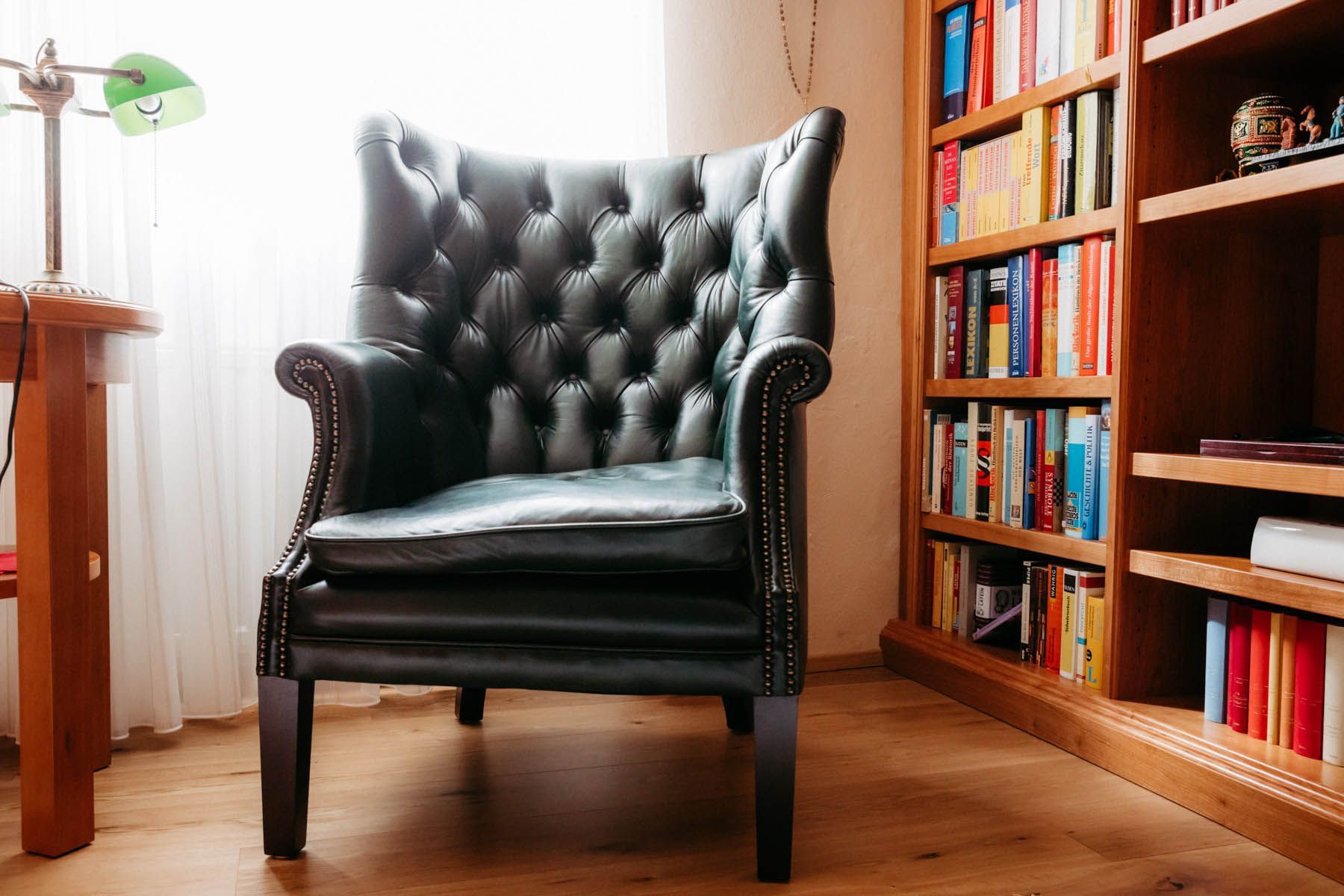 Schwarzer Ledersessel mit Polsterknöpfen neben einem Bücherregal, beleuchtet von einer Schreibtischlampe.