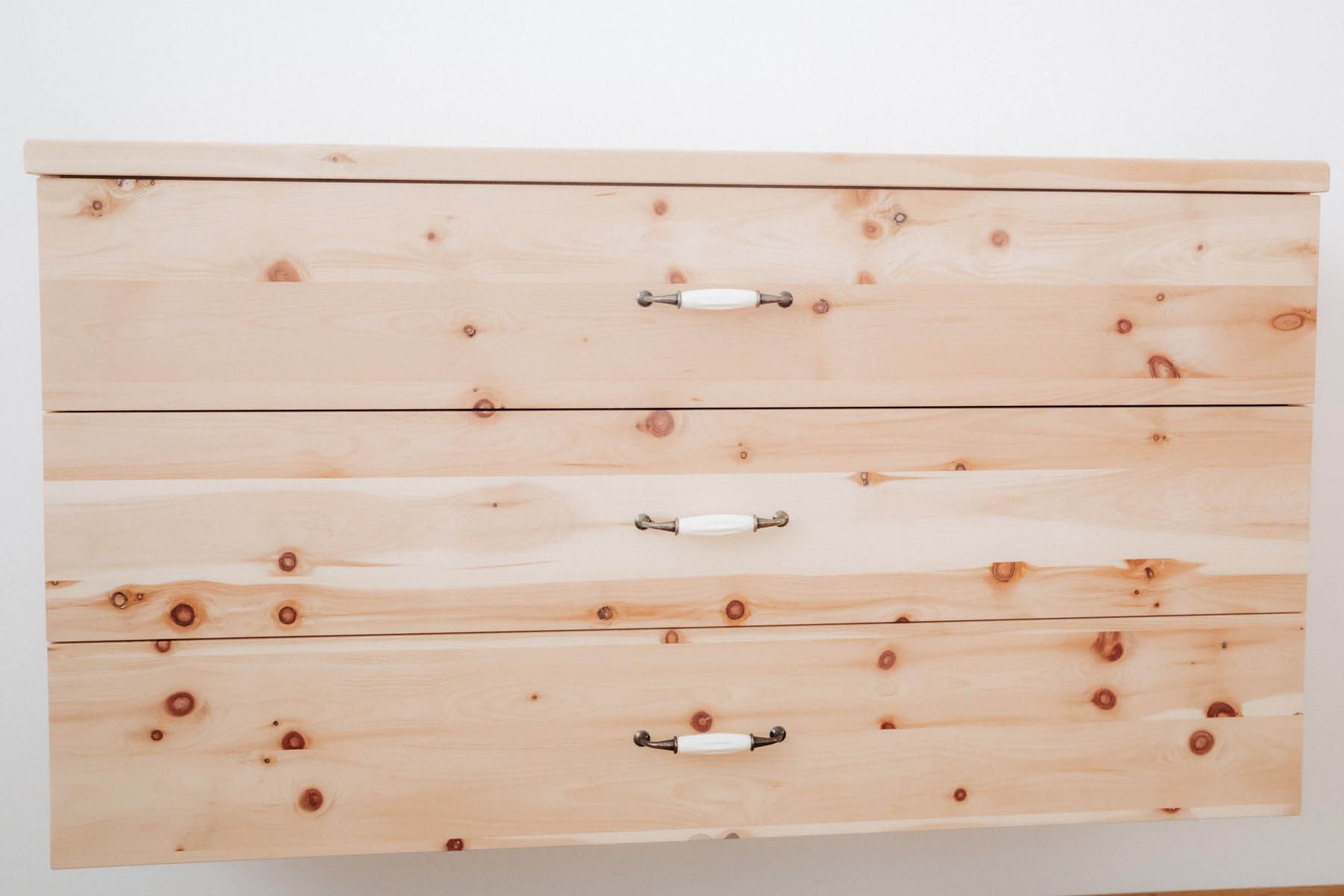 Holzkommode mit drei Schubladen und weißen Griffen, an einer weißen Wand montiert.