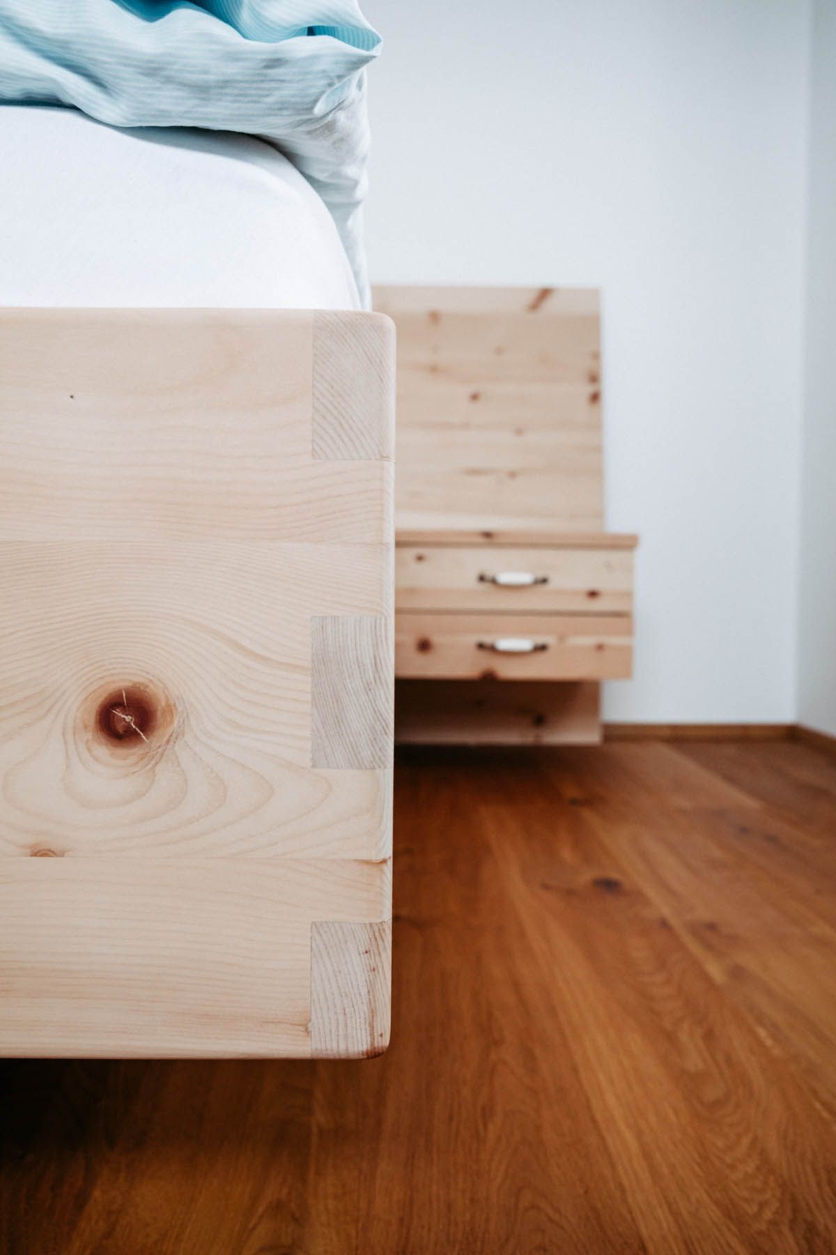 Holzbettrahmen mit Schwalbenschwanzverbindungen, neben einem passenden Nachttisch auf Holzboden.