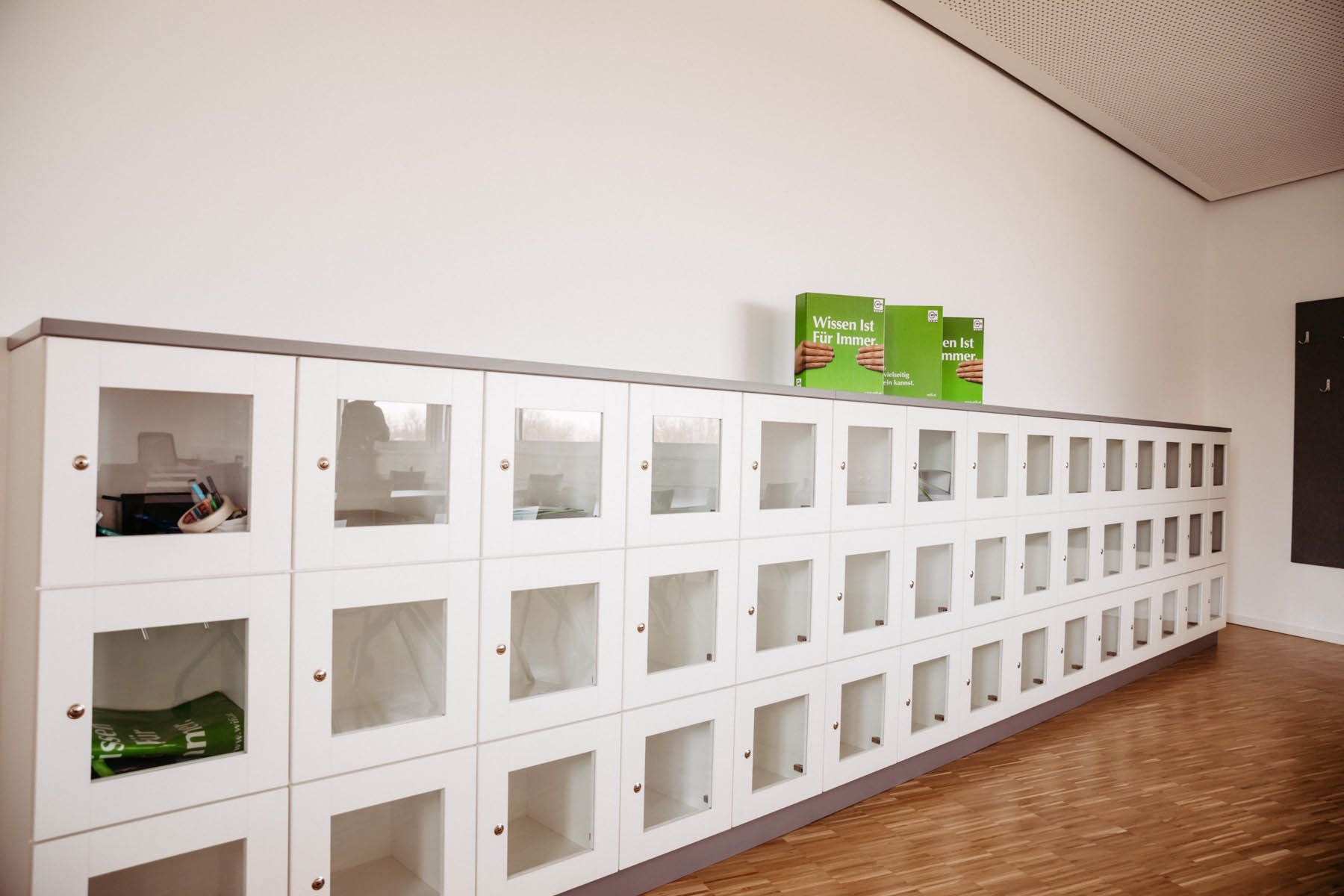 An einer Wand stehen weiße Schließfächer mit Glastüren, in denen verschiedene Gegenstände aufbewahrt werden. Oben drauf stehen drei grüne Kisten.