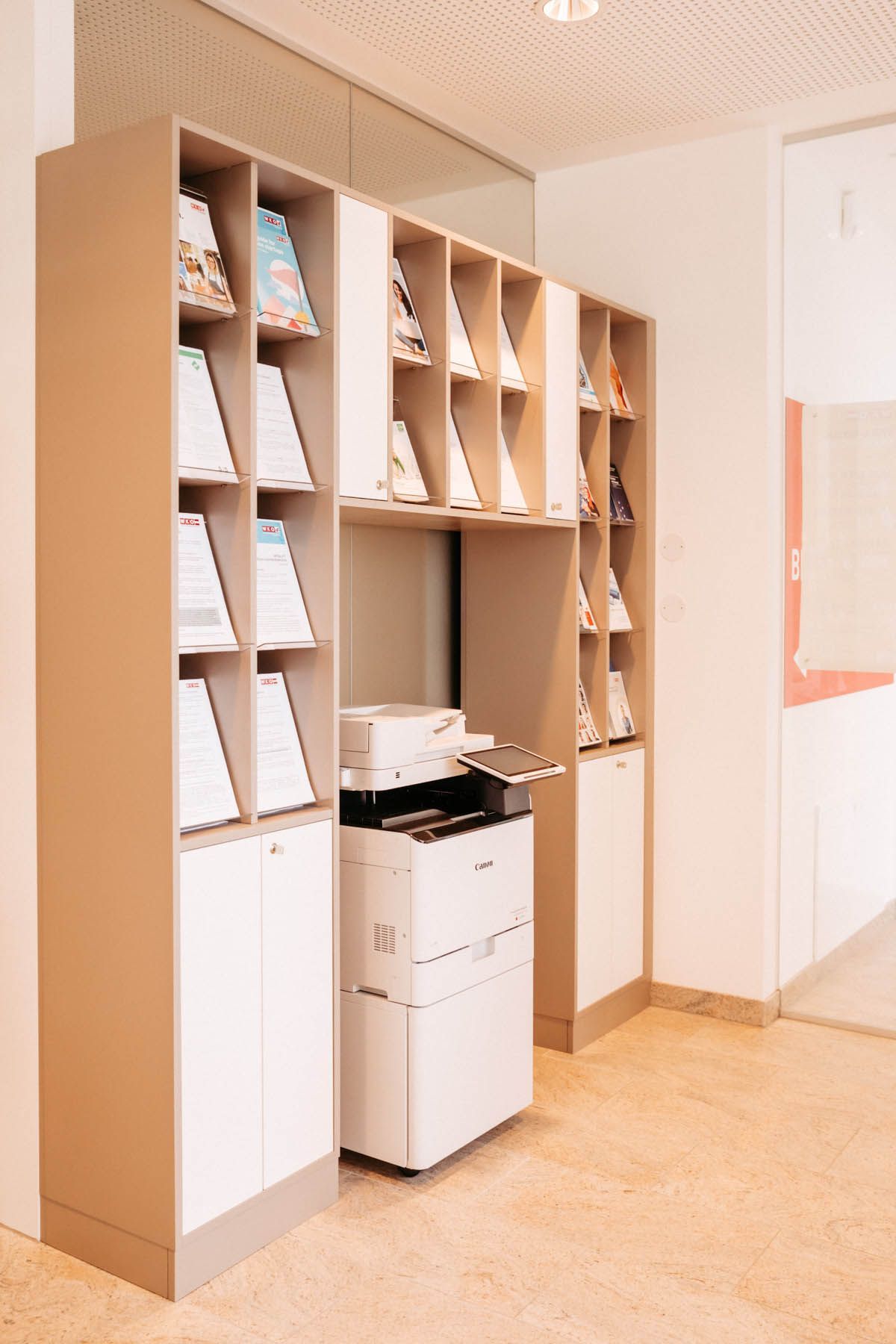 Holzregal mit Broschüren und Drucker. Neutrales Farbschema.