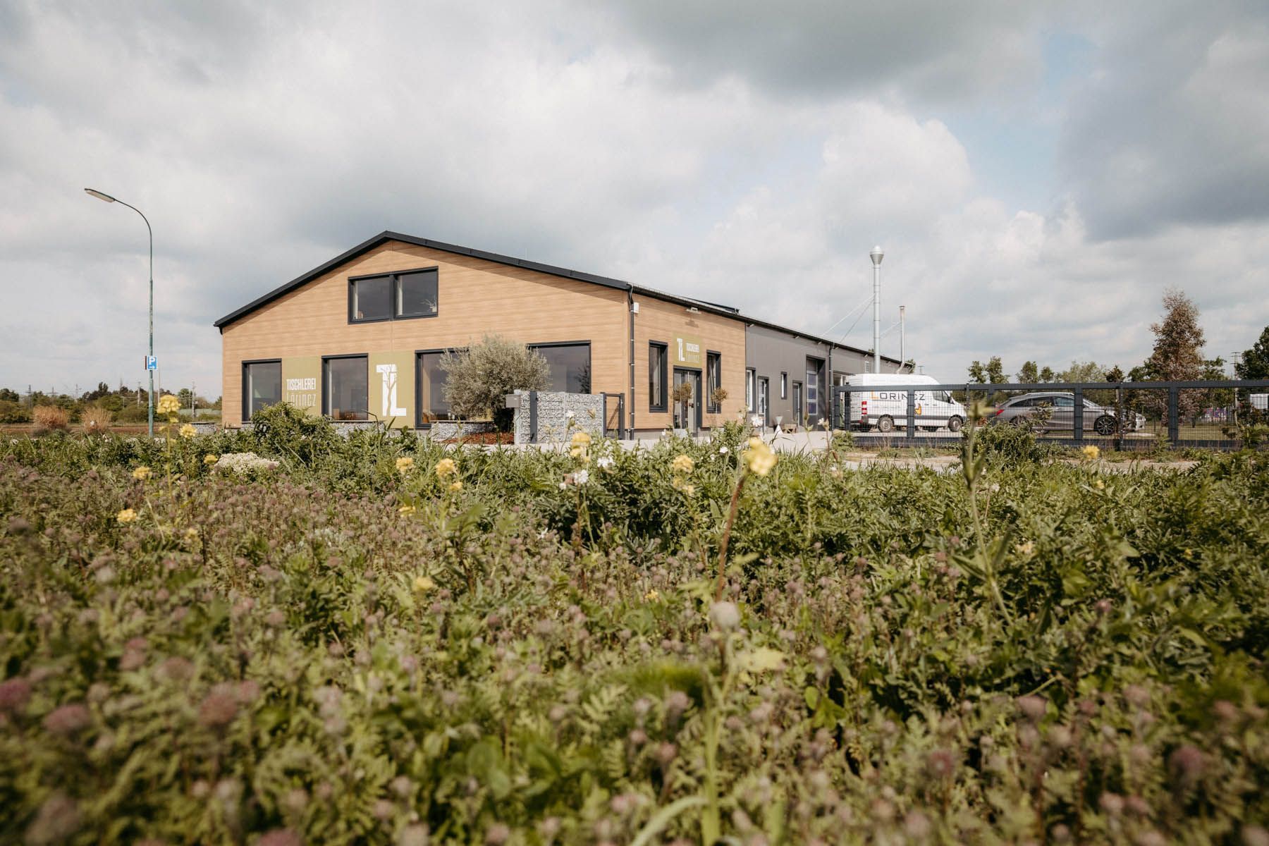 Backsteingebäude mit dunklem Dach auf einem Feld mit Wildblumen unter einem bewölkten Himmel.