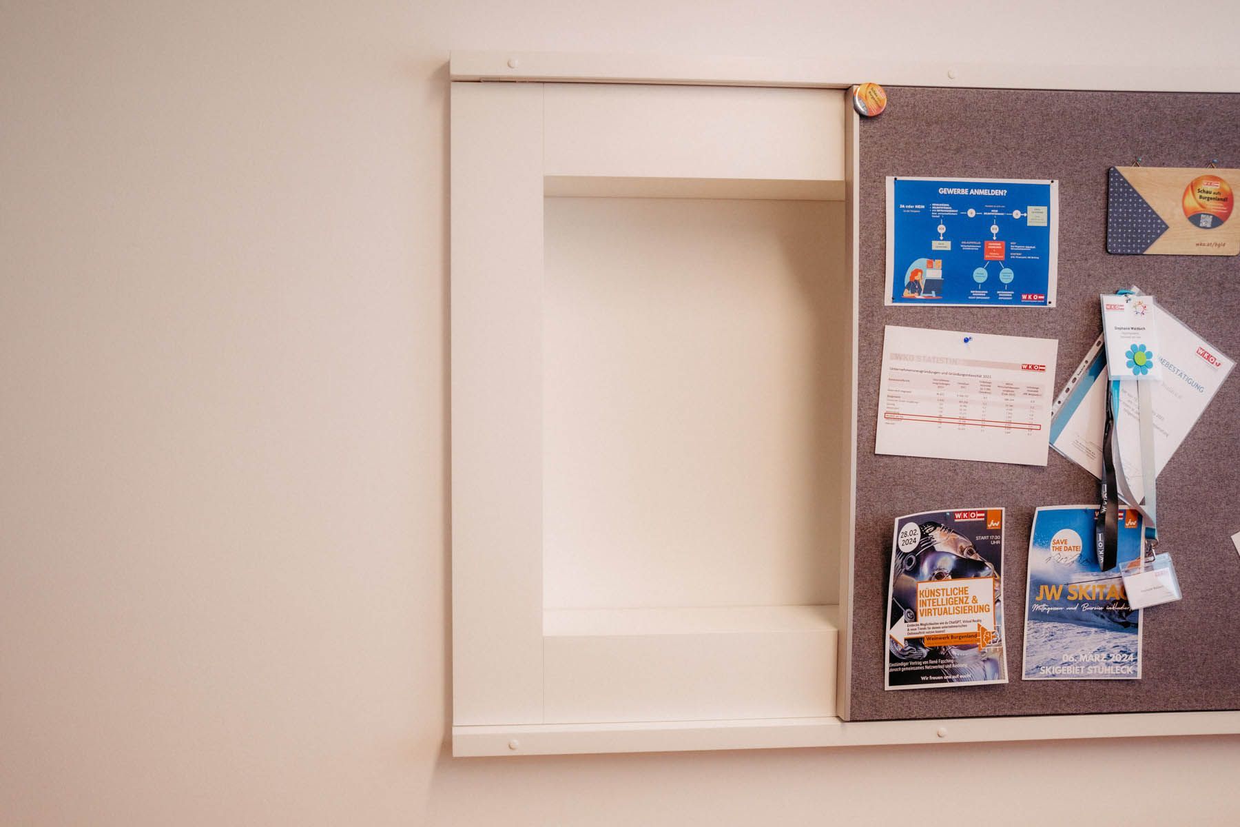 Weiße Wand mit eingebauter rechteckiger Nische neben einem mit Papieren und Stecknadeln bedeckten schwarzen Brett.