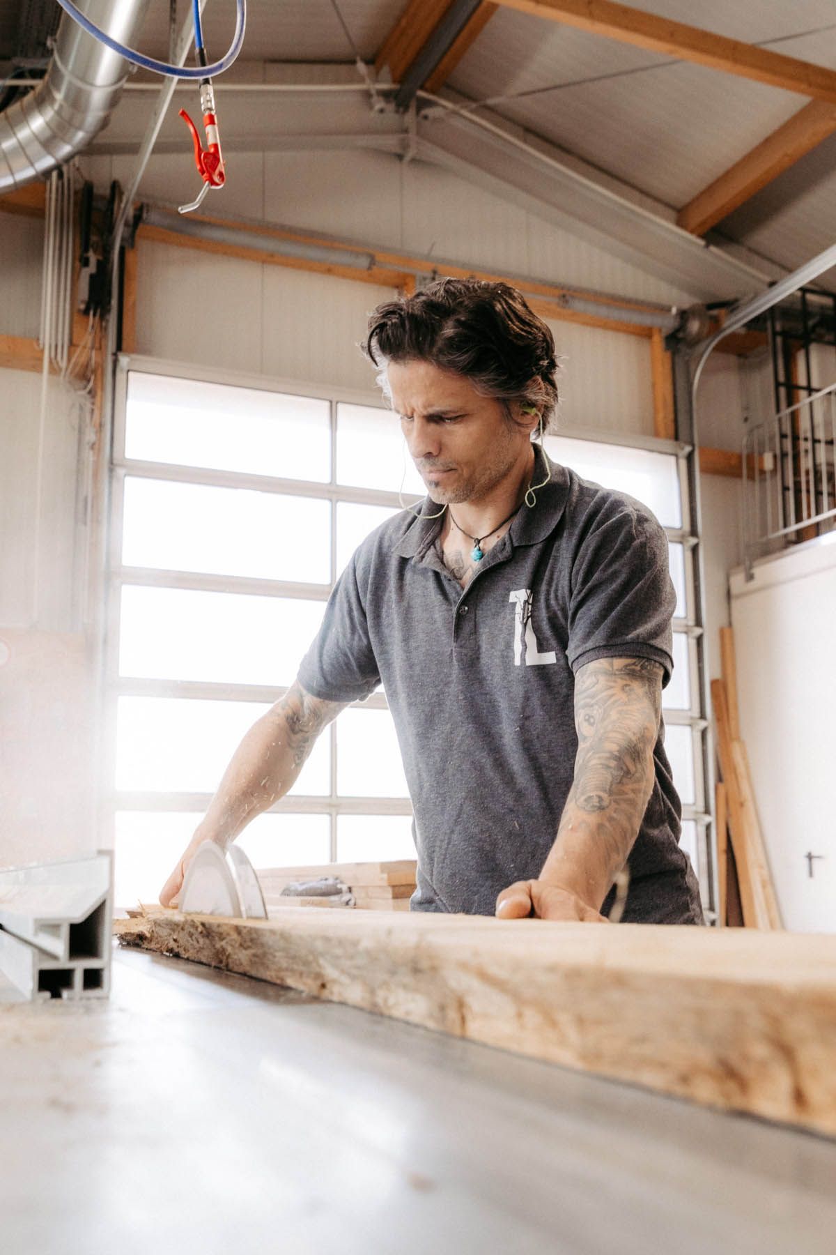 Ein Mann in einer Werkstatt sägt mit einer Säge ein Holzbrett. Er trägt ein T-Shirt und hat Tattoos auf dem Arm.