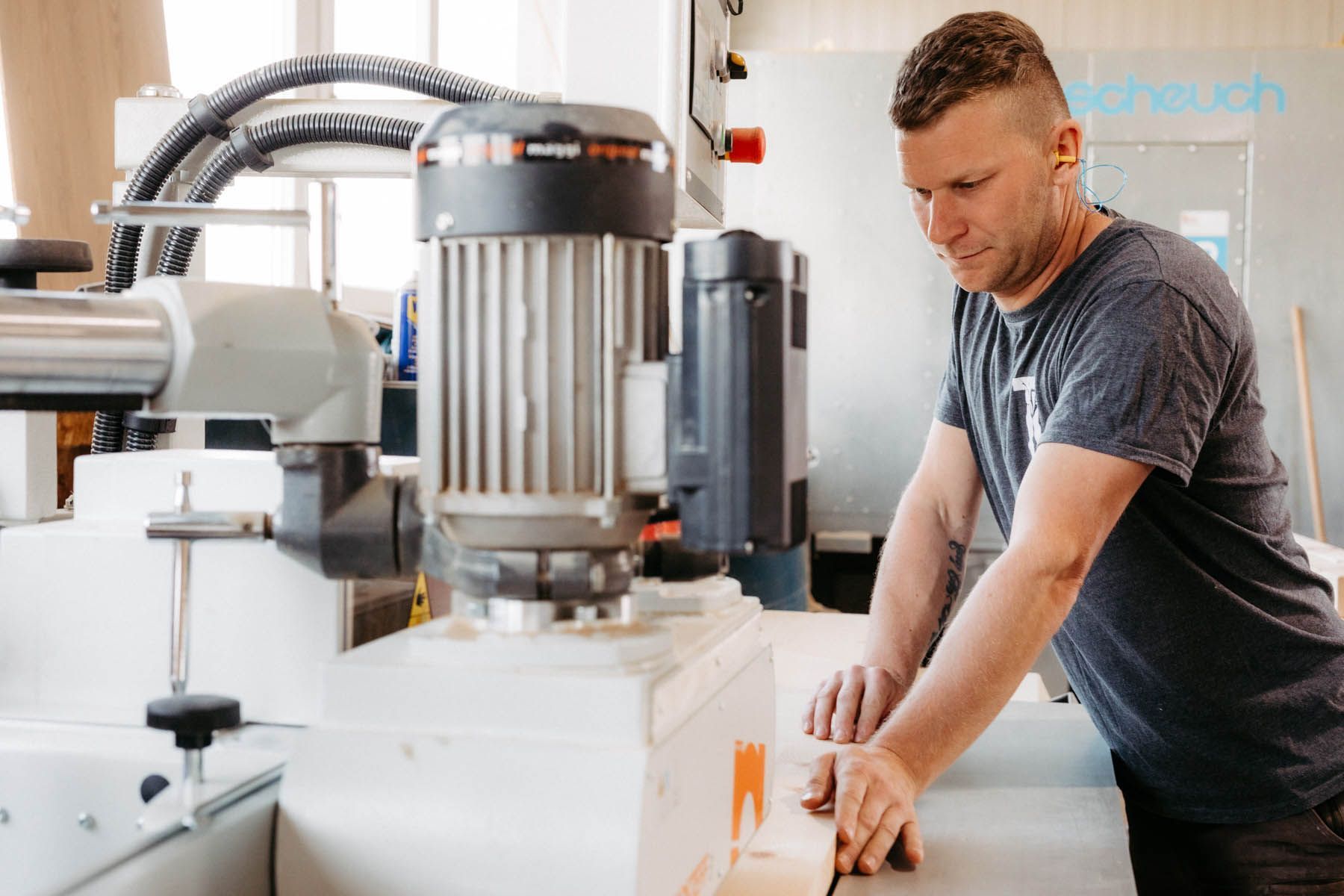 Mann arbeitet in einer Werkstatt an einer Holzmaschine. Er trägt ein graues Hemd und konzentriert sich auf das Holzstück.