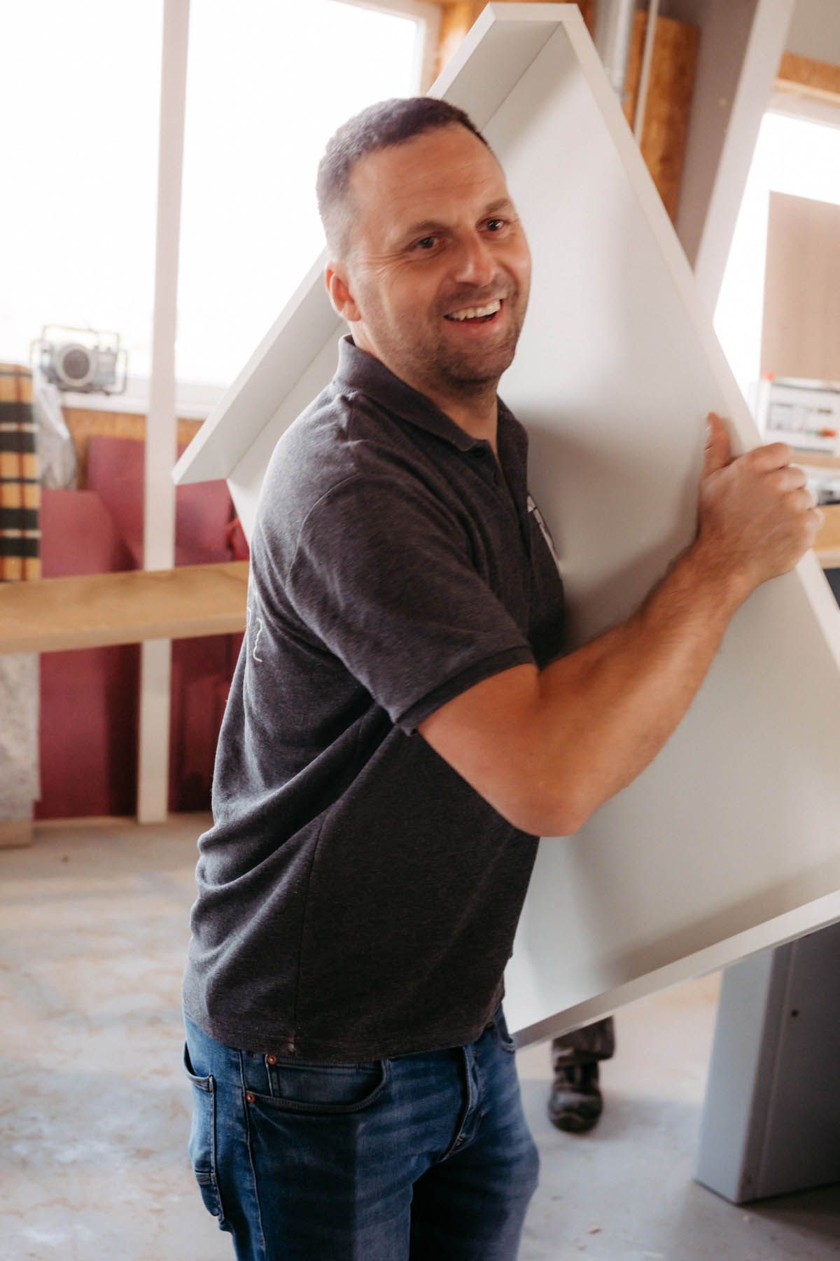 Lächelnder Mann in grauem Hemd und Jeans, der einen großen weißen Gegenstand in einer Werkstatt trägt.