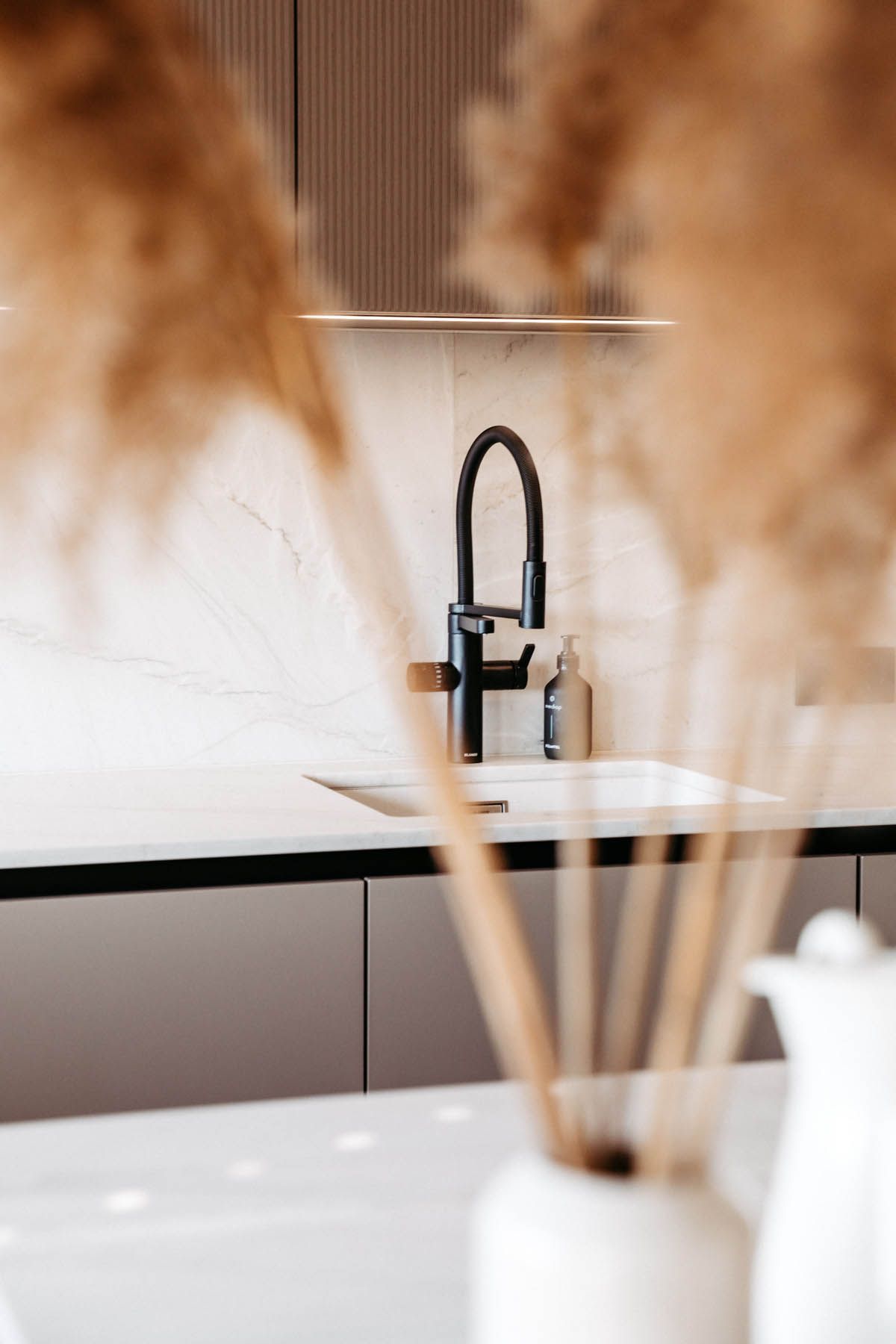 Schwarzer Küchenhahn vor einer hellen Marmorrückwand, durch eine Vase mit getrockneten Pflanzen fokussiert.