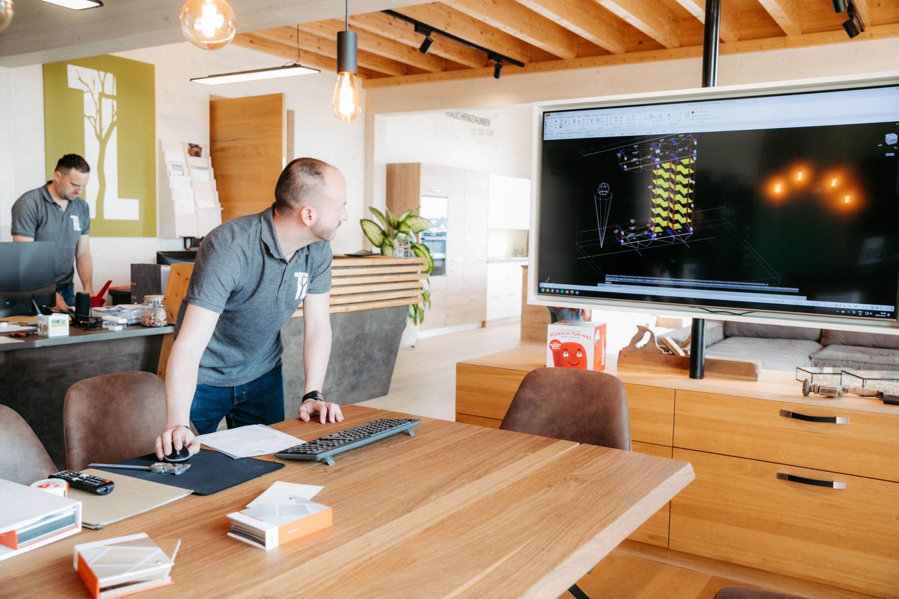 Zwei Männer in einem Büro schauen auf einen großen Bildschirm, auf dem ein Design angezeigt wird. Holztisch und -schränke, Lampen.