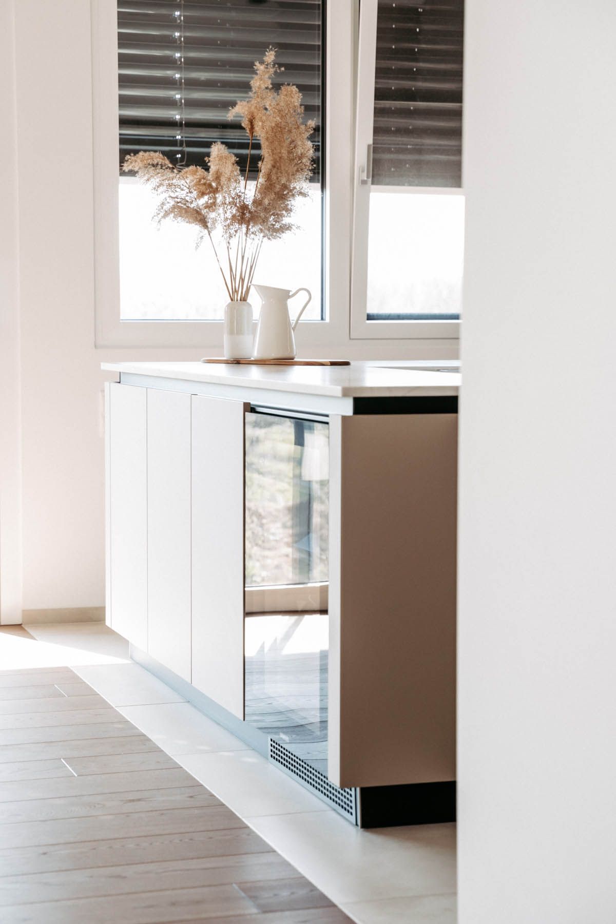 Weiße, moderne Küche mit eingebautem Weinkühler, natürlichem Licht durch das Fenster und dekorativen Trockenblumen.