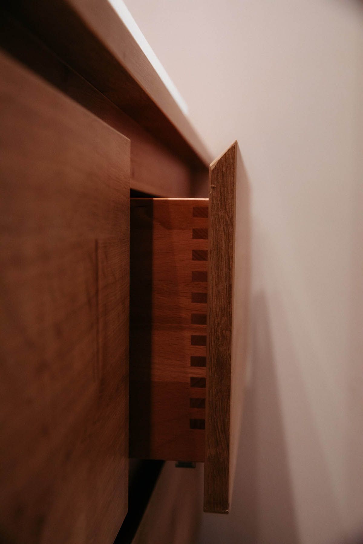Teilweise geöffnete Holzschublade mit sichtbaren Schwalbenschwanzverbindungen. Braunes Holz vor einer weißen Wand.