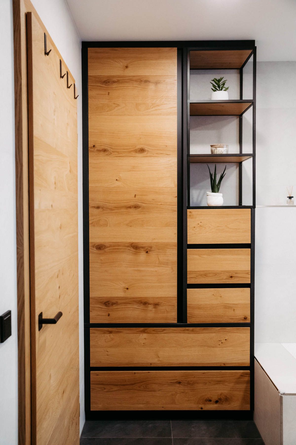 Holzschrank mit schwarzem Rahmen in einem Badezimmer, inklusive Regalen, Schubladen und einer Tür.