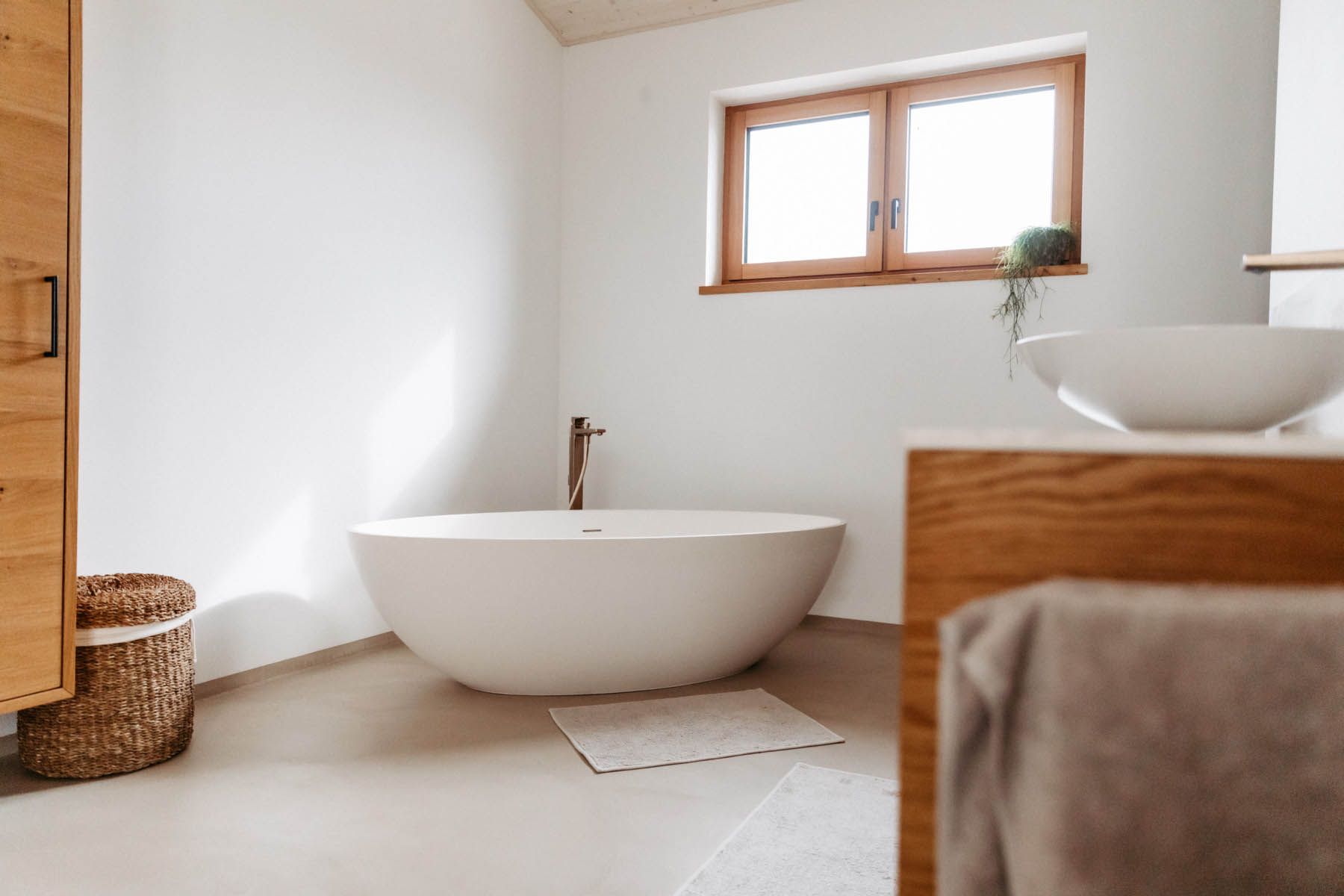 Badezimmer mit einer weißen ovalen Badewanne, hellen Holzakzenten und einem Fenster mit Tageslicht.