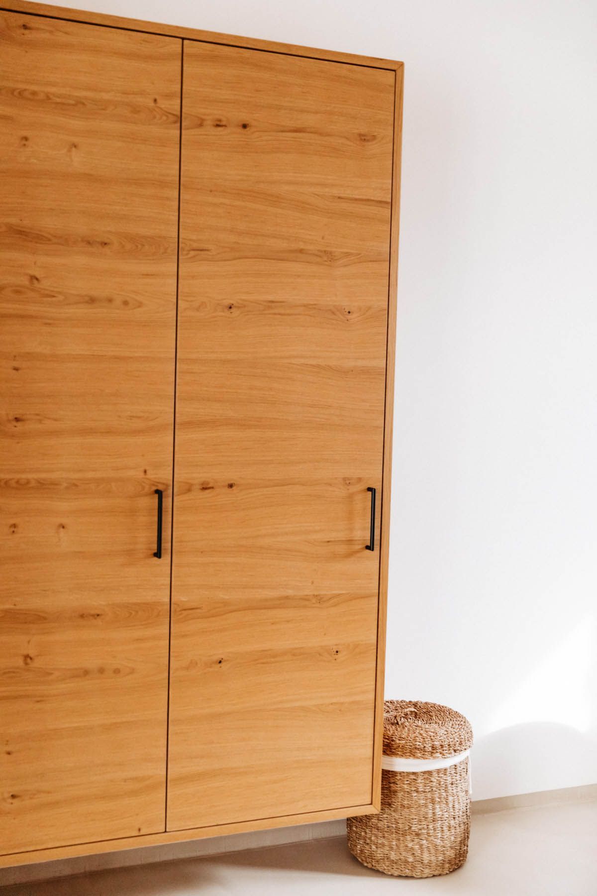 Holzschrank mit schwarzen Griffen vor einer weißen Wand. Darunter steht ein geflochtener Korb.