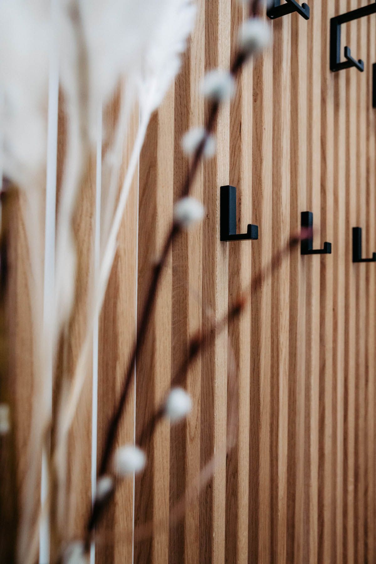 Holzpaneelwand mit schwarzen Haken, Zweig mit weißen Knospen und verschwommenen Blüten.